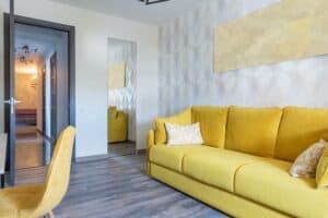 Mayorż&oacute;łta sofa w nowoczesnym salonie z geometryczną tapetą i drewnianymi podłogami.